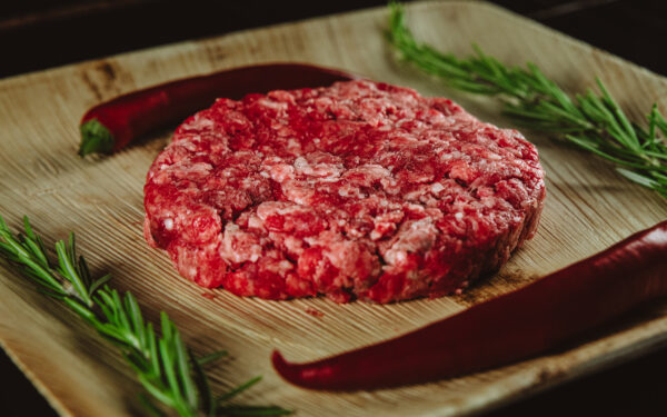 Surowy burger wołowy na desce drewnianej z papryką czerwoną jako dekoracją. Alesmakosz.pl