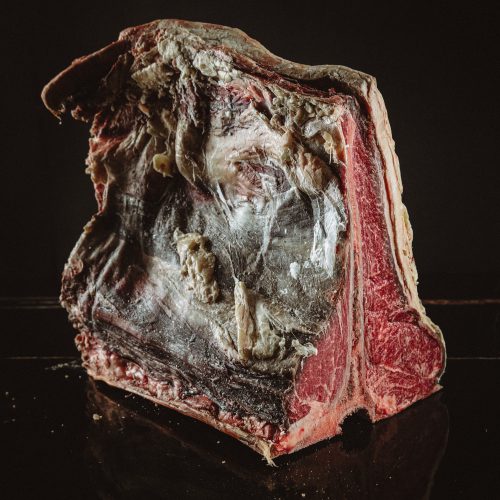 Fiorentina z mięsa wołowego z kością i okrywą sezonowana na sucho pochodząca z Polski. Stojąca na stole z czarnego granitu na czarnym tle.