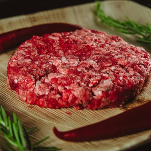 Surowy burger wołowy na desce drewnianej z papryką czerwoną jako dekoracją. Alesmakosz.pl